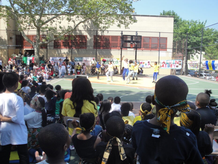 Students and their families gather outside on school basketball court to watch young dancers present a dance routine.