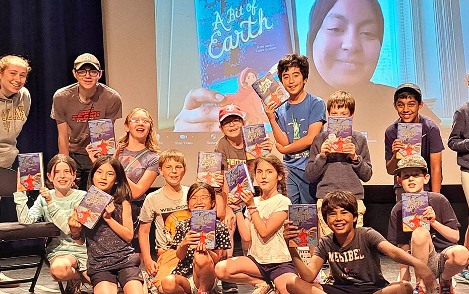 Campers smile and hold up copies of a book while gathered on a stage. The book's author, Karuna Riazi, poses the same way, projected on a screen behind them.