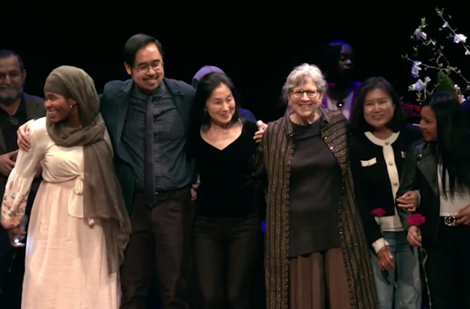 A diverse group of adults, who are All Write! program participants and staff, stand on a stage arm-in-arm and smile at audience - this links to a video.