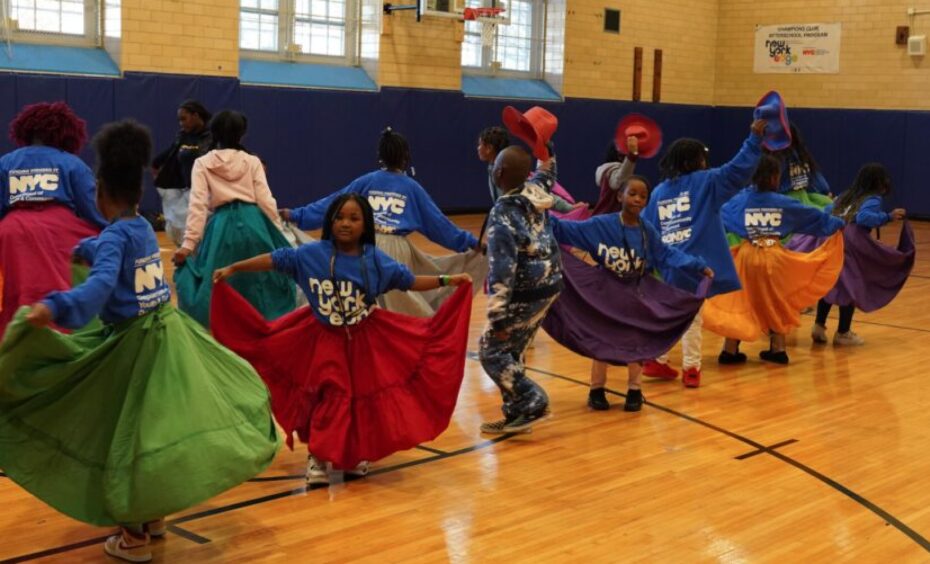 Children in Global Arts Mexican Folk Dance workshop smile and dance in a school gymnasium wearing brightly colored skirts and hats.
