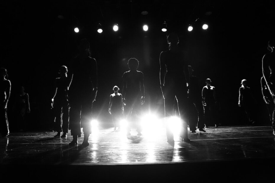 Black and white photo. Twelve dancers pose as statues on stage as they are uplit with bright stage lights obscuring their faces.