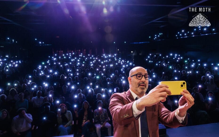 Host Amir Baghdadchi with his back to the audience takes a phone selfie. All of the audience have their phone flashlights on in the dark theater.