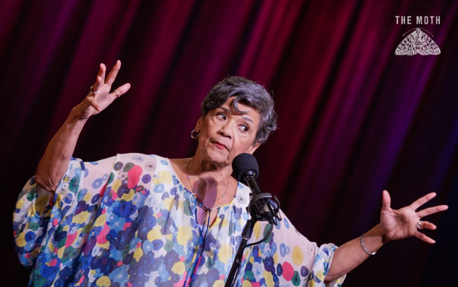 Storyteller Sonia Manzano stands in front of a red curtain. She has her hands outstretched and is speaking to the audience.