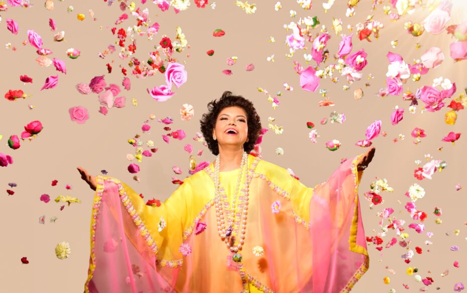 Chandrika Tandon appears wearing a flowing yellow and pink rob, surrounded by falling pink petals on a tan background.