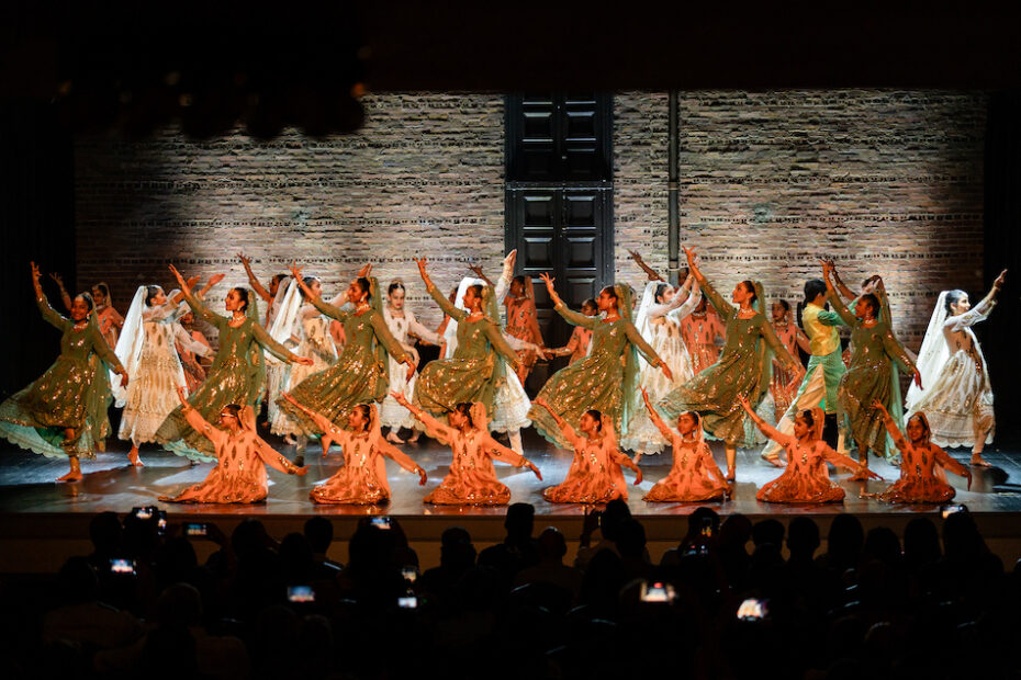 Three lines of dancers pose on stage. They are in three different levels and lit with orange and white lights.