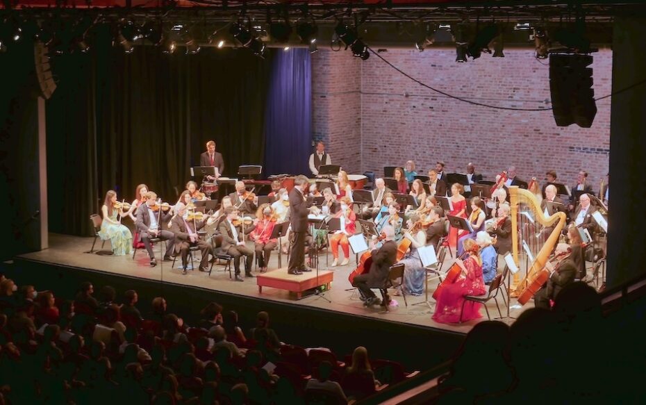A full symphony orchestra appears on a lit stage with brick back wall. The photo is taken from an upper balcony.
