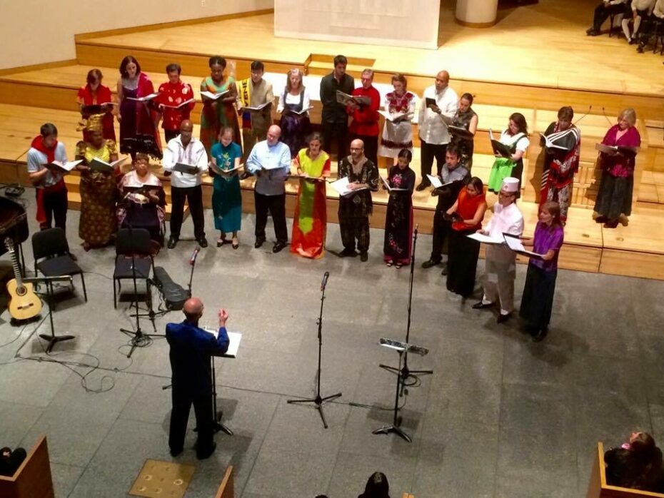 A group of 15 vocalists stand in front of a conductor mid-song. They wear traditional outfits from Nigeria, China, Japan, Hungary, and others.