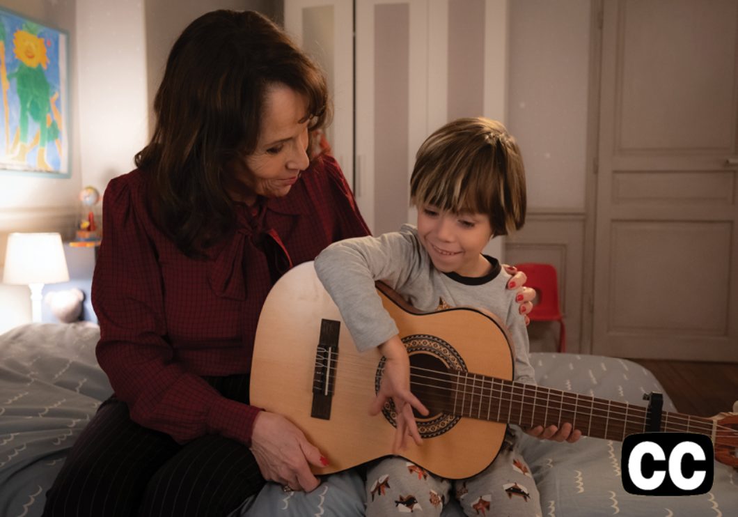 A grandmother and a young boy with a guitar, sitting on the child's bed.