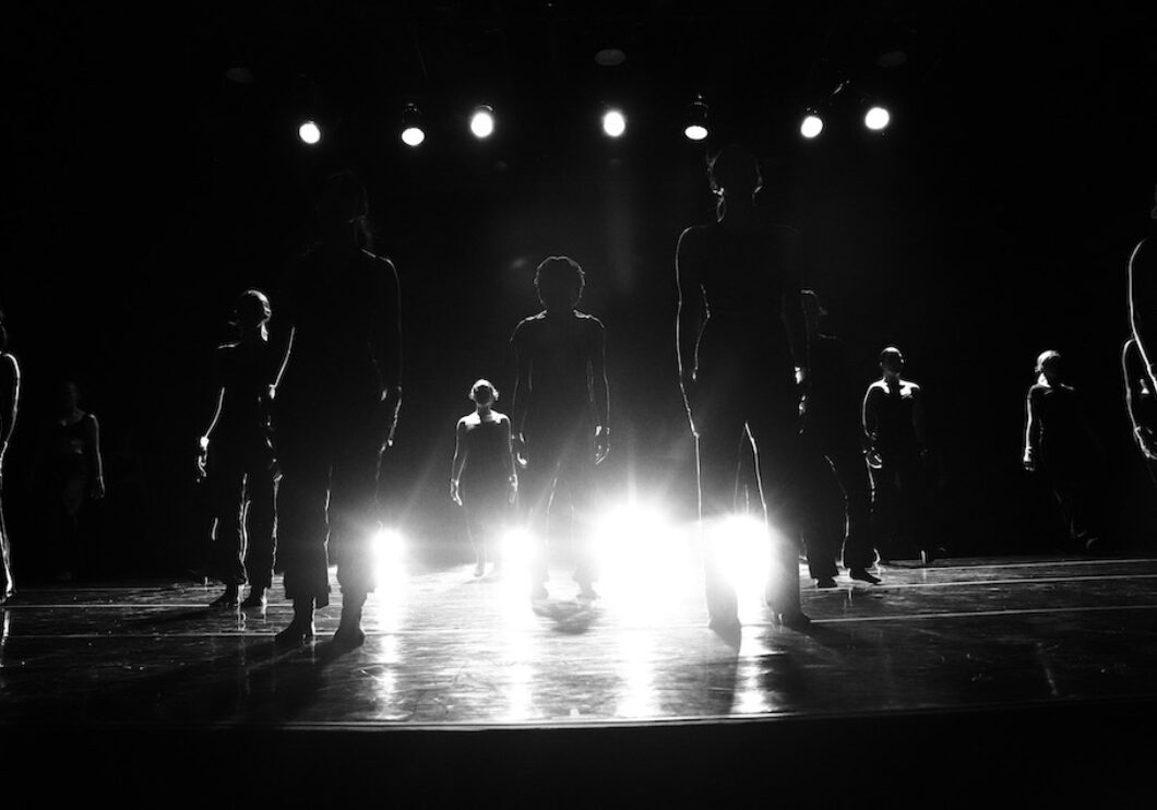 Black and white photo. Twelve dancers pose as statues on stage as they are uplit with bright stage lights obscuring their faces.