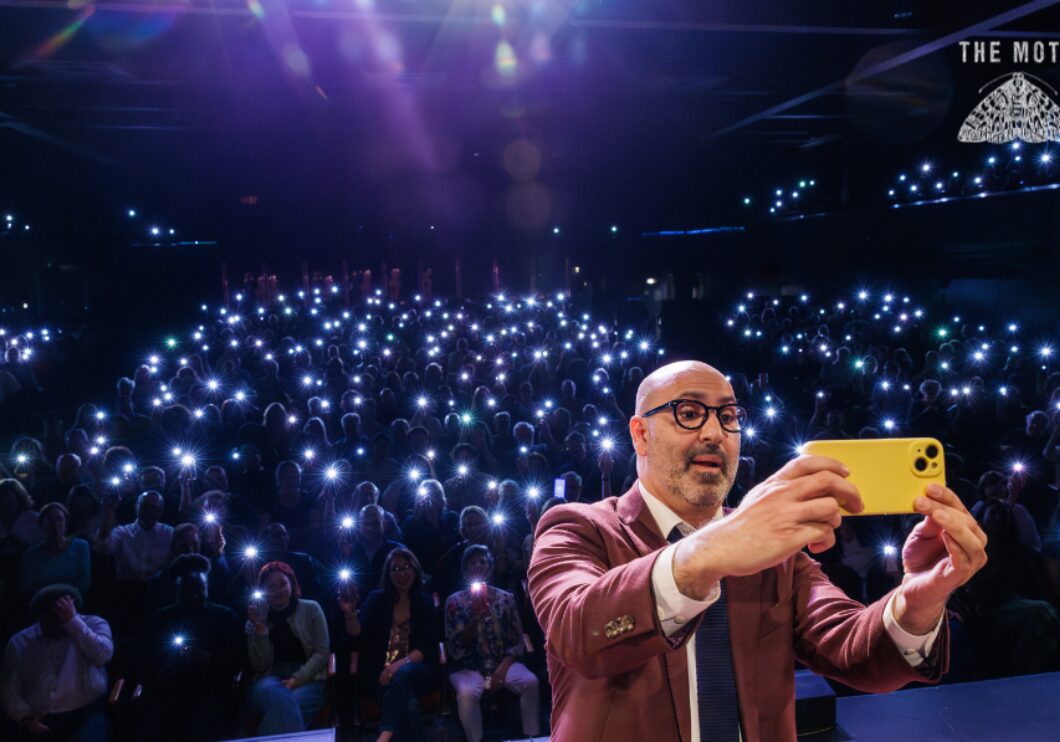 Host Amir Baghdadchi with his back to the audience takes a phone selfie. All of the audience have their phone flashlights on in the dark theater.