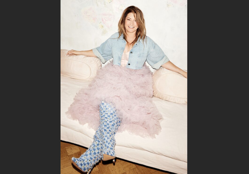 Pianist Helen Jane Long sits with her legs crossed on a white couch. She is wearing a denim coverall suit with pink tutu and smiling at the camera.