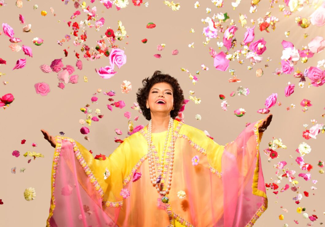 Chandrika Tandon appears wearing a flowing yellow and pink rob, surrounded by falling pink petals on a tan background.