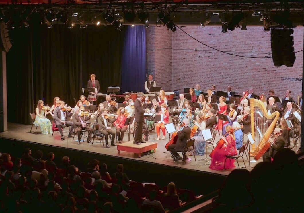 A full symphony orchestra appears on a lit stage with brick back wall. The photo is taken from an upper balcony.