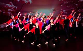 An ensemble of dancers strike a pose on stage. They wear black jumpsuits with red overcoats, and are lit by stage lights from above.