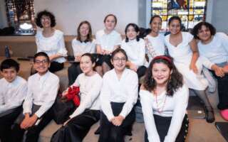 Twelve members of a choir sit on two benches., six in front of the other. They are all smiling and wearing white shirts.