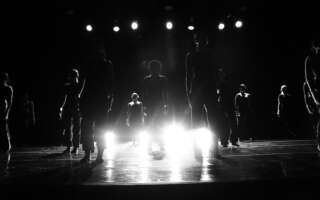 Black and white photo. Twelve dancers pose as statues on stage as they are uplit with bright stage lights obscuring their faces.