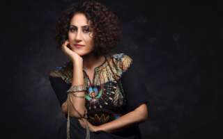 Portrait of artist Aynur against a black background. She is leaning her chin on her right hand and wearing decorative blue jewelry.