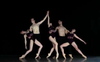 A group of modern dancers pose on stage against a black curtain. They all wear black.
