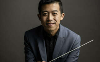Conductor Alex Wen headshot. He is holding a baton over a dark gray background.