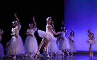 A group of ballet dancers in white tutus pose mid-dance in front of a purple lit backdrop.
