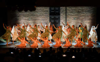 Three lines of dancers pose on stage. They are in three different levels and lit with orange and white lights.