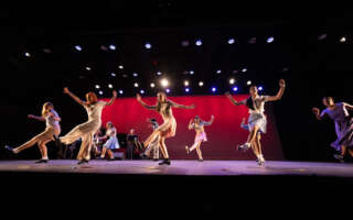 Two spaced out lines of dancers tap on a fully lit stage. A piano and jazz quartet are behind them.