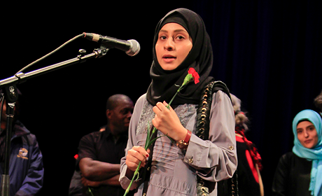 A young woman wearing a hijab stands on stage near a microphone.